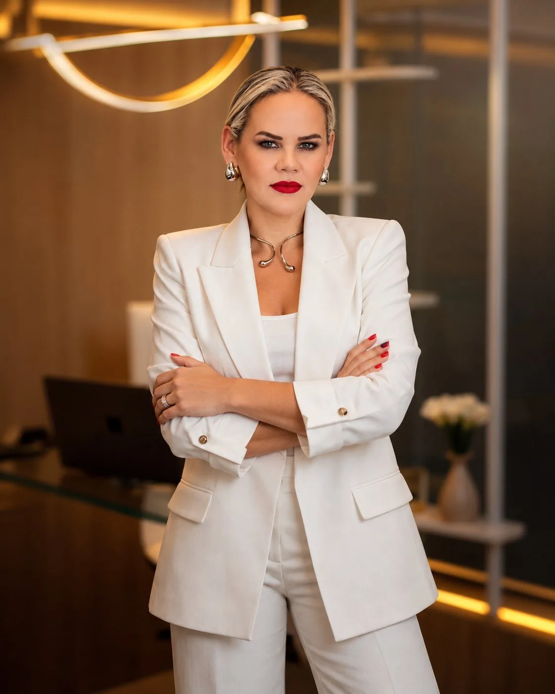 Paula Teixeira de terno branco, braços cruzados, em um escritório com luz quente